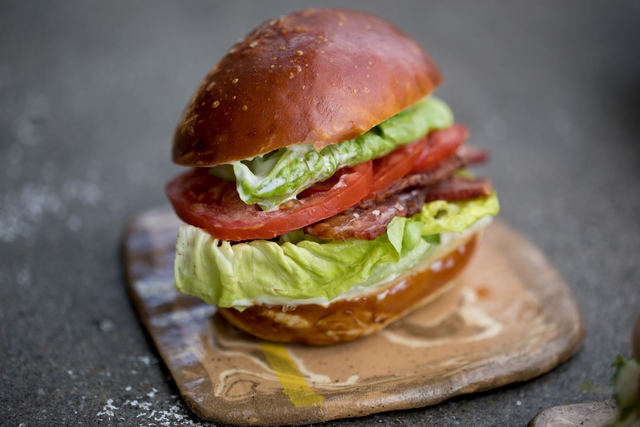 A sandwich with crispy bacon, fresh lettuce, and sliced tomato is placed on a square rustic wooden board.